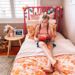 A Dawn calendar and reminder Clock shown on bedside table as child with a disability (Cerebral Palsy) relaxes and reads a book. The girl has blonde hair and pigtail plaits and wears a pink shirt and denim shorts. There are bright flowers on her bedsheets.