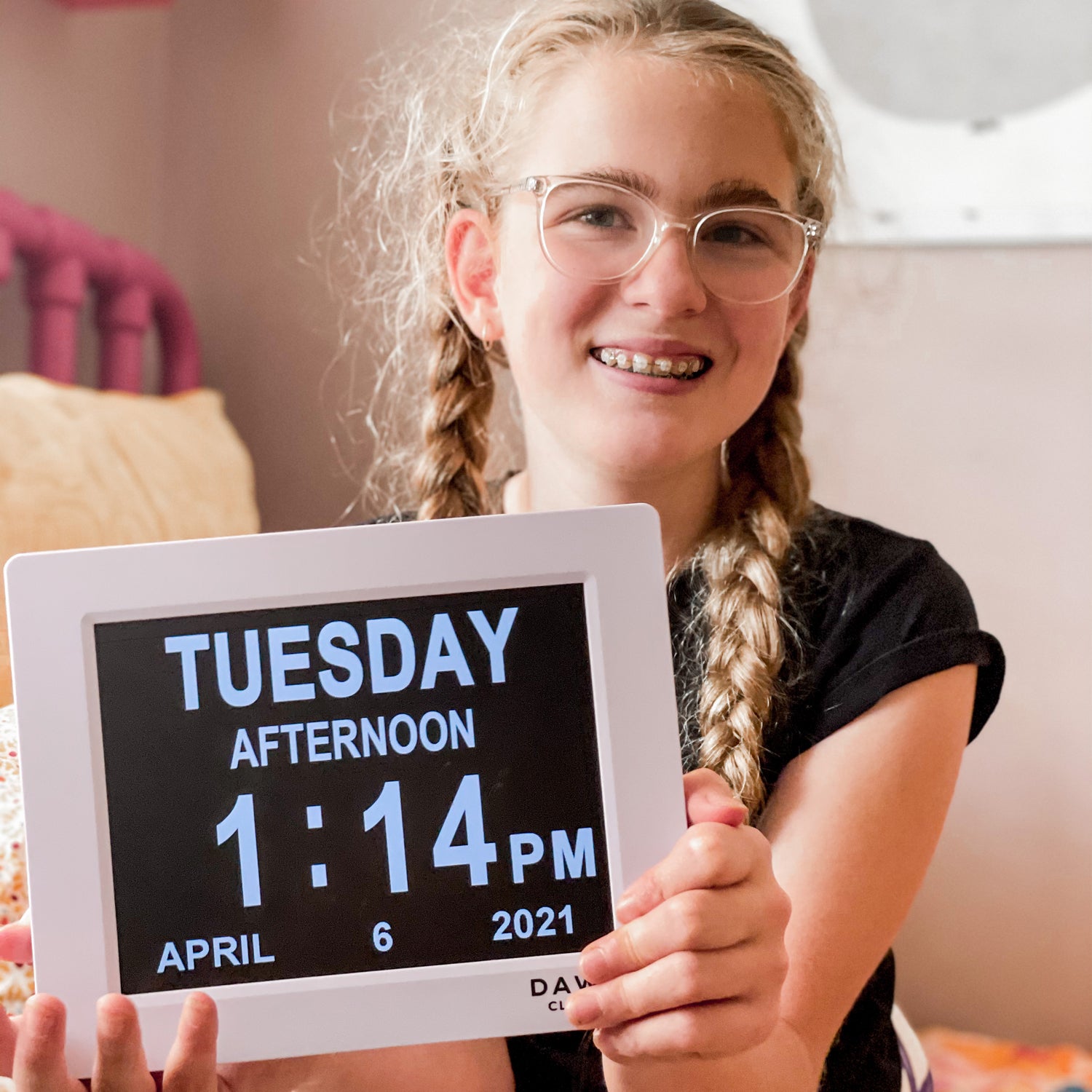 A girl holds a Dawn Clock - Calendar Day Date Time Digital reminder clock. The date says Tuesday afternoon 1:14pm April 6 2021/. The frame is white and the writing is white on a black background. Emily has glasses and plaits. She is wearing a black T-shirt and grey tracksuits pants while sitting on a bed. Emily is very happy holding her Dawn Clock. NDIS Cerebral Palsy ADD ADHD Autism ASD Neurodiversity Brain Injury Memory Loss Dementia Alzheimer's Disease Disability Alarm Gift Plan Manager Plan Managed.